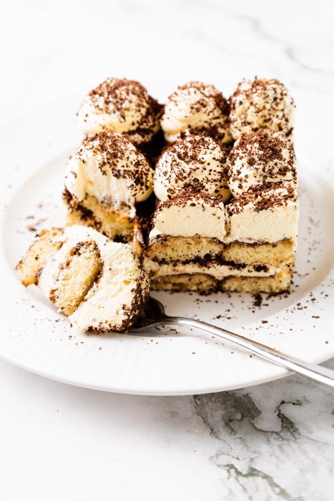 A slice of tiramisu with layers of creamy filling and cocoa is served on a white plate, with a fork holding a piece and cocoa powder sprinkled on top—perfect to enjoy alongside a cup of hot chocolate.