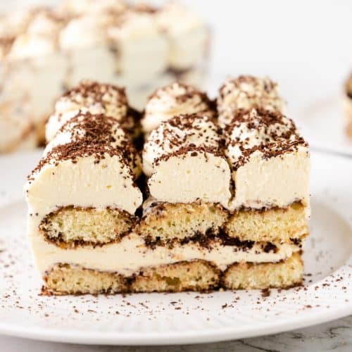 A slice of tiramisu on a white plate, showing layers of creamy filling, coffee-soaked ladyfingers, and a dusting of chocolate shavings—perfect to enjoy alongside a cup of hot chocolate. More slices are visible in the background.