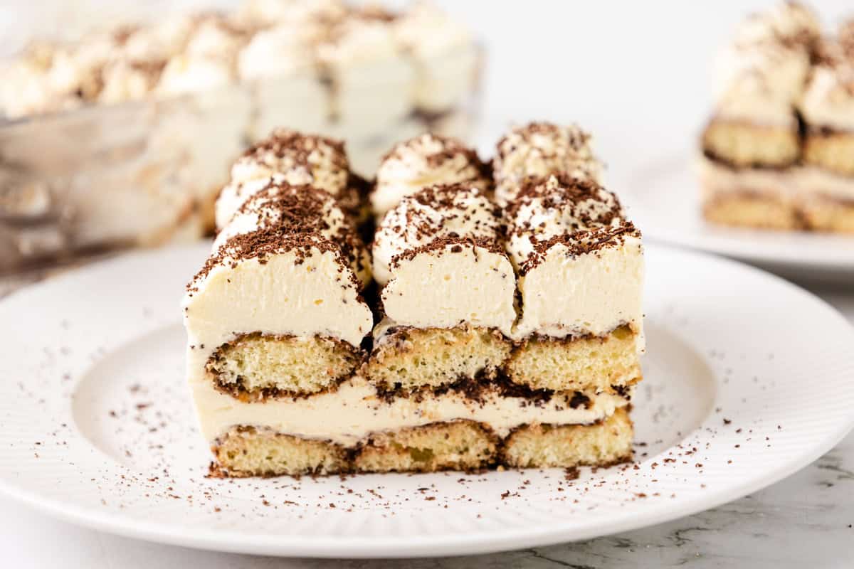 A slice of tiramisu on a white plate, showing layers of creamy filling, coffee-soaked ladyfingers, and a dusting of chocolate shavings—perfect to enjoy alongside a cup of hot chocolate. More slices are visible in the background.