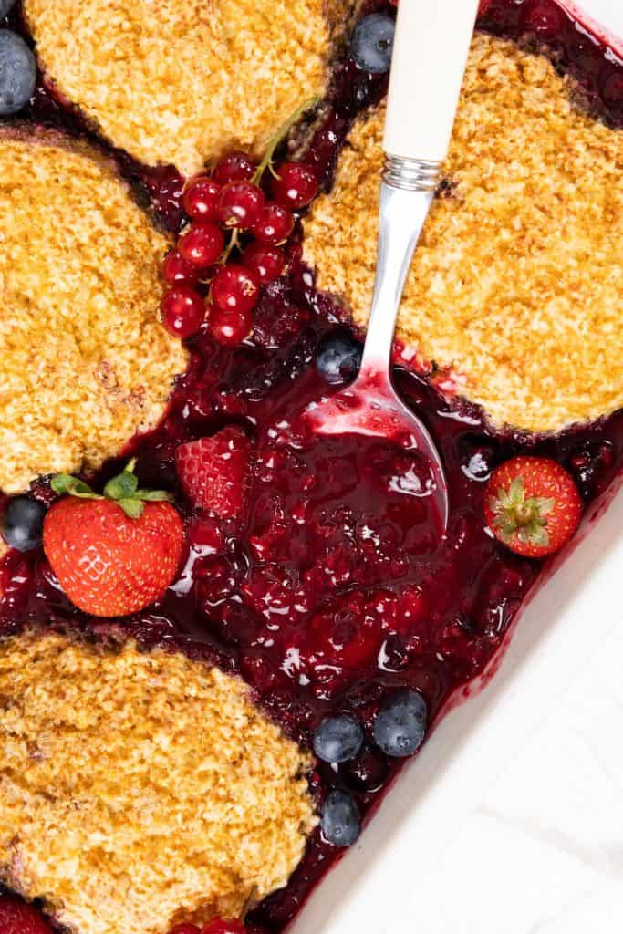 A close-up of a luscious Berry Cobbler with golden biscuits, juicy berry filling, strawberries, blueberries, red currants, and a spoon scooping out the dessert.