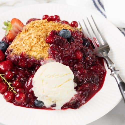 A plate with berry cobbler, a scoop of vanilla ice cream, and a fork. Fresh berries and sauce are spread around the dessert, with a striped napkin beside the plate.