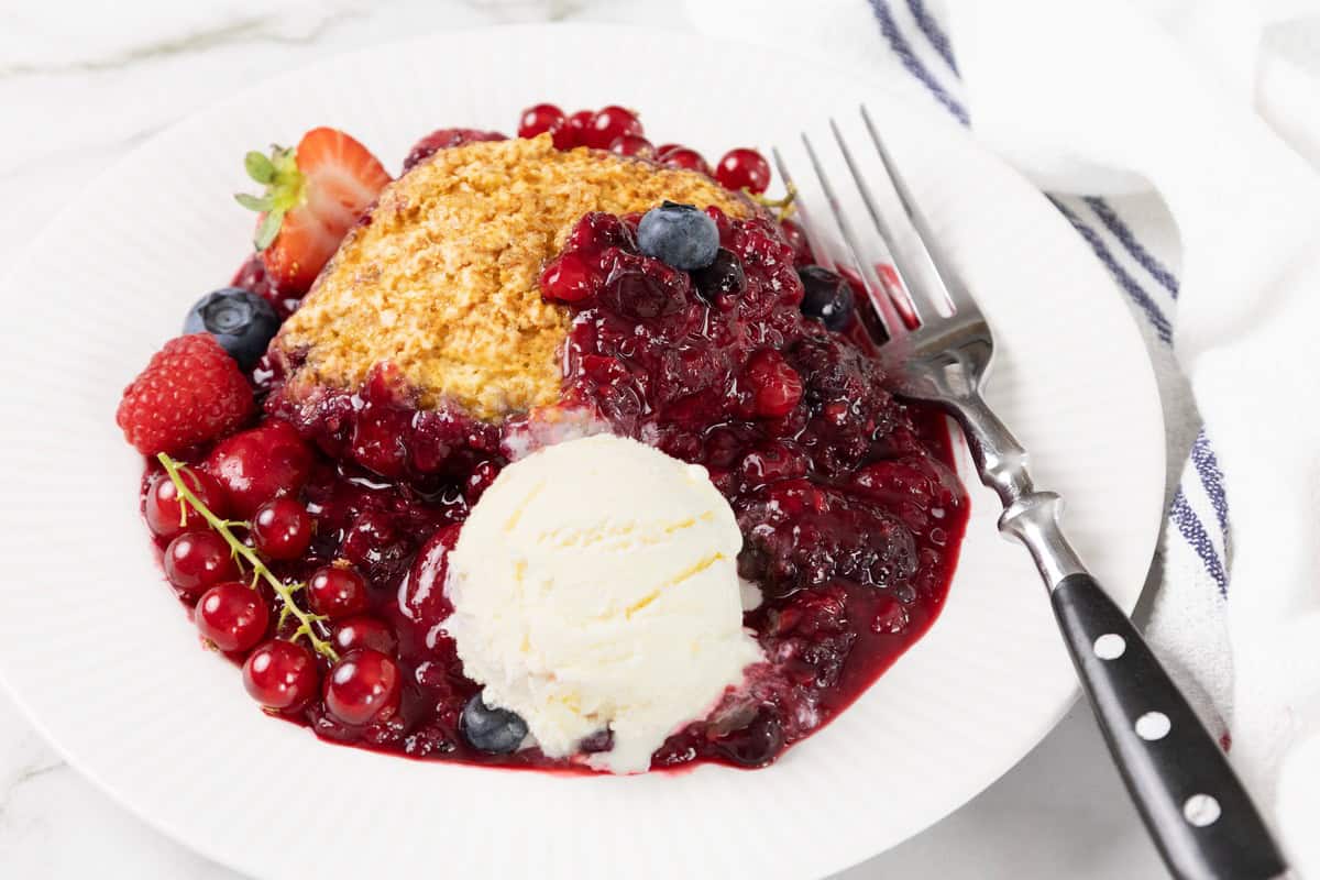 A plate with berry cobbler, a scoop of vanilla ice cream, and a fork. Fresh berries and sauce are spread around the dessert, with a striped napkin beside the plate.