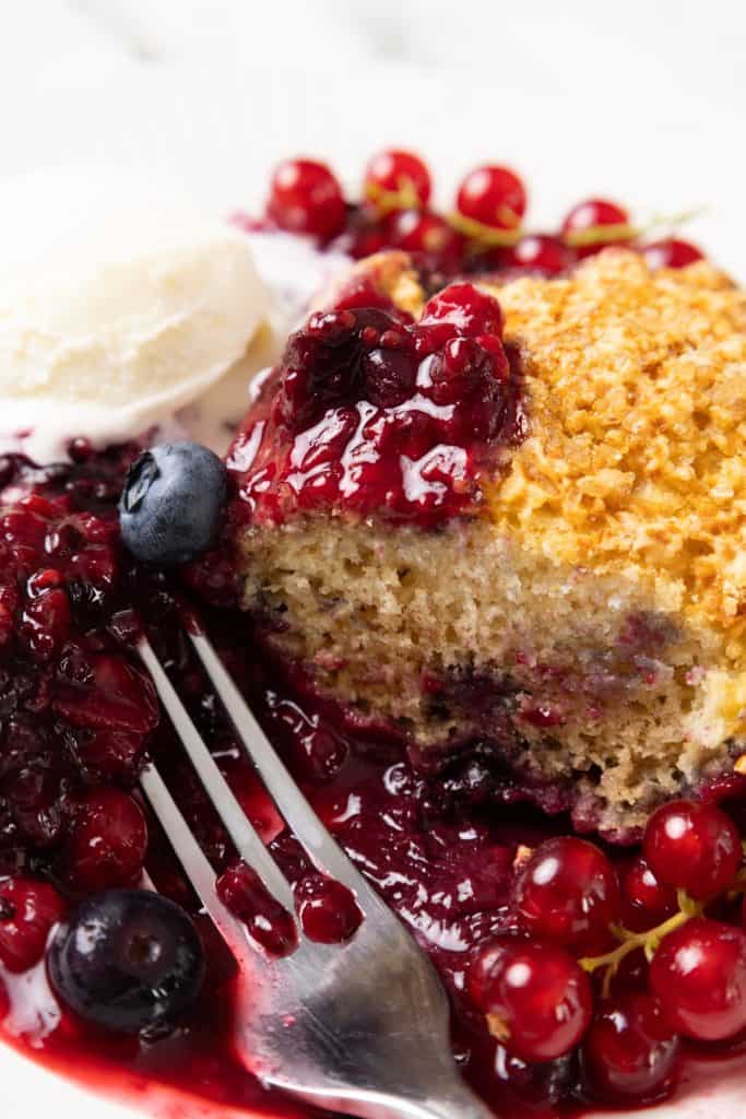A close-up of Berry Cobbler with golden crust, surrounded by mixed berries and sauce, served with a scoop of vanilla ice cream on a white plate, and a fork resting beside it.