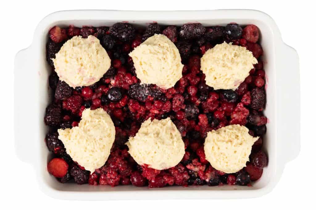A white baking dish filled with mixed berries and topped with six scoops of biscuit dough, ready to be baked into a delicious Berry Cobbler.