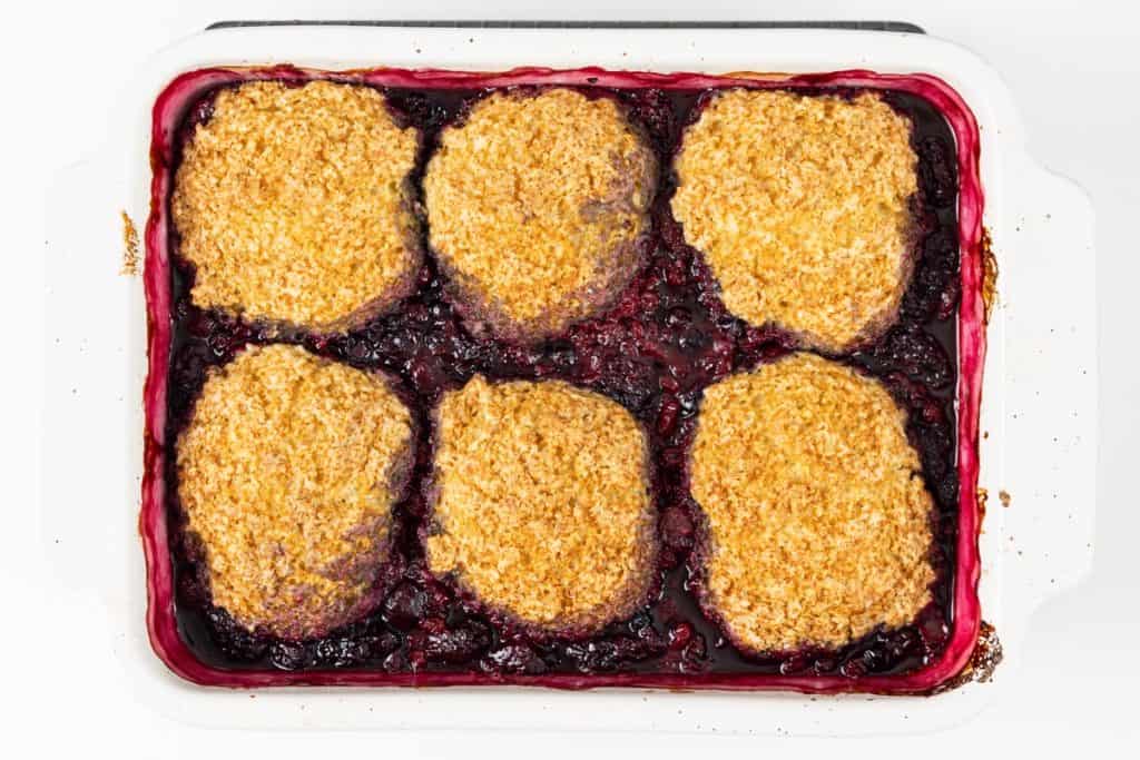 A Berry Cobbler baked in a white dish, topped with six golden-brown biscuit pieces, with dark berry filling bubbling around the edges.