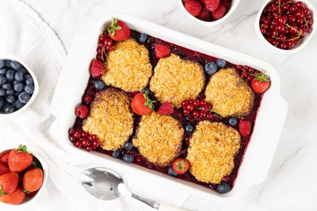 A Berry Cobbler is beautifully presented in a white dish, topped with six golden biscuits and surrounded by fresh strawberries, blueberries, and red currants in small bowls on a marble surface.