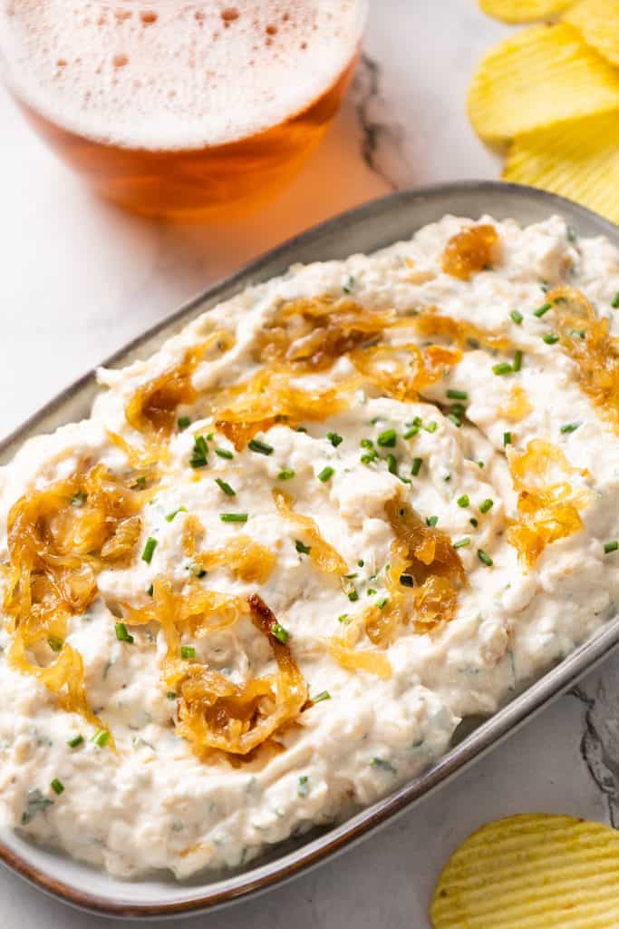An oval dish of creamy Caramelized Onion Dip topped with golden caramelized onions and chopped chives, surrounded by ridged potato chips. A glass of amber-colored drink with foam sits in the background on a marble surface.