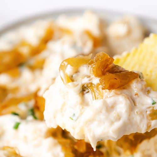 A hand with red nail polish holds a ridged potato chip topped with creamy Caramelized Onion Dip and caramelized onions, with a bowl of the onion dip in the background.