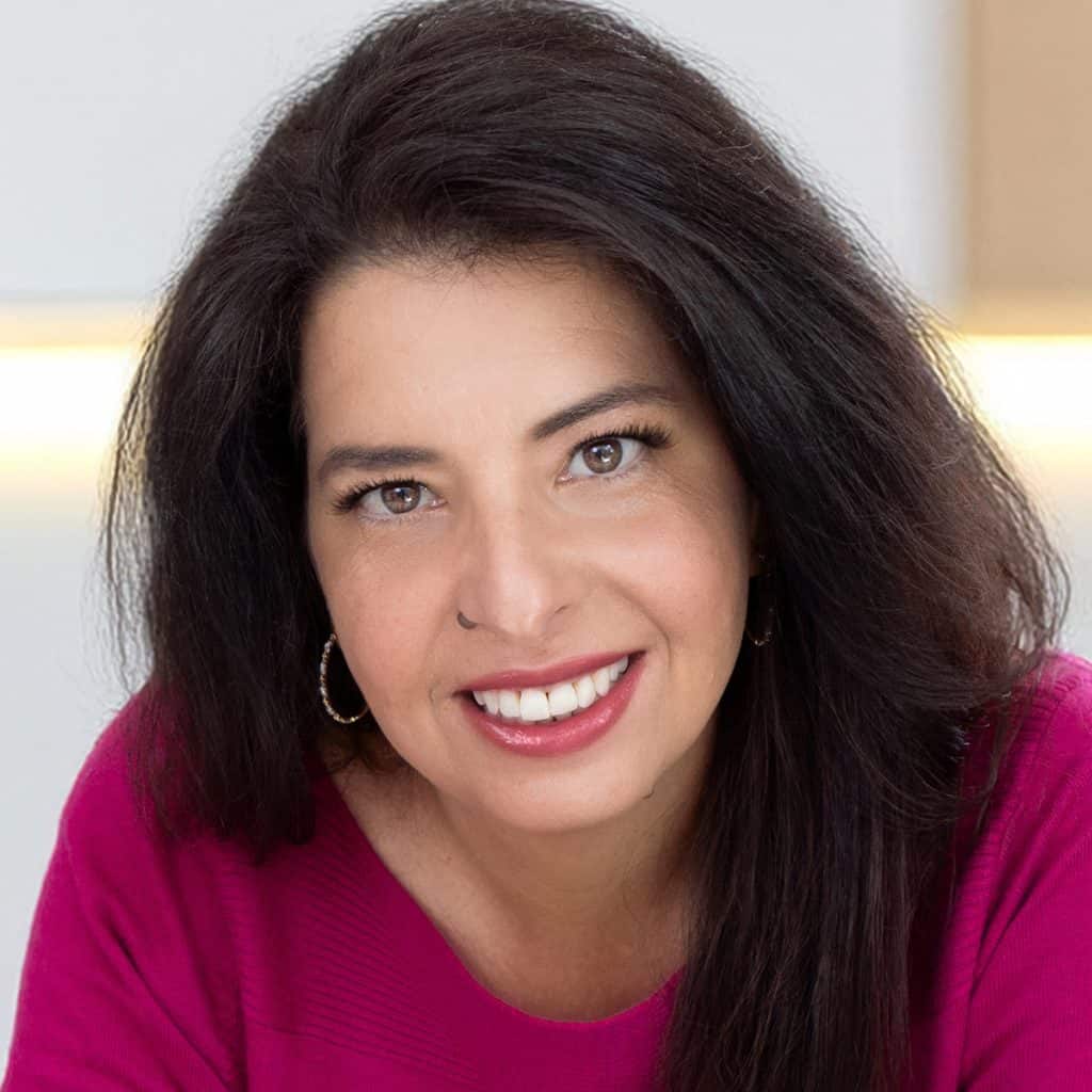 About Erren Hart: A woman with long dark hair smiles warmly at the camera. She is wearing a magenta top and hoop earrings, with a neutral background behind her.