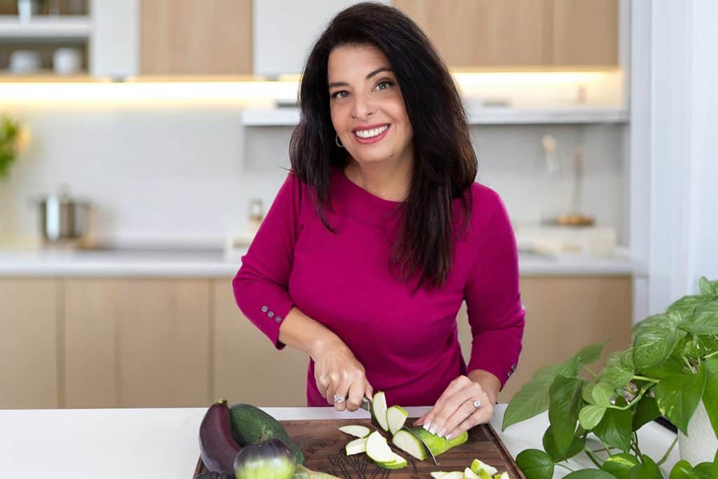 Erren Hart with long dark hair, wearing a magenta top, smiles while slicing zucchini on a wooden cutting board in a modern kitchen with light cabinets and green plants.