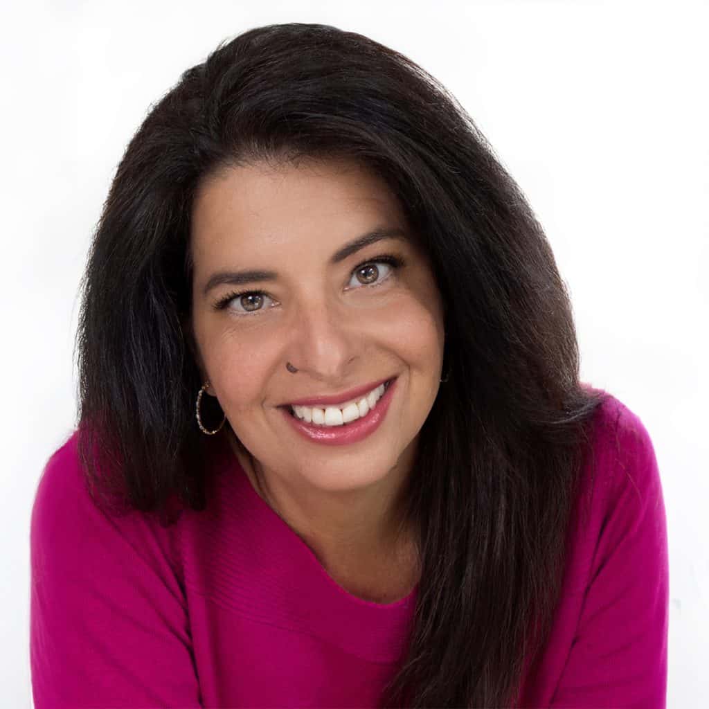 Erren Hart smiling woman with long dark hair wearing a bright pink sweater and gold hoop earrings, photographed against a white background—capturing the warmth and approachable style that’s all About Erren Hart.