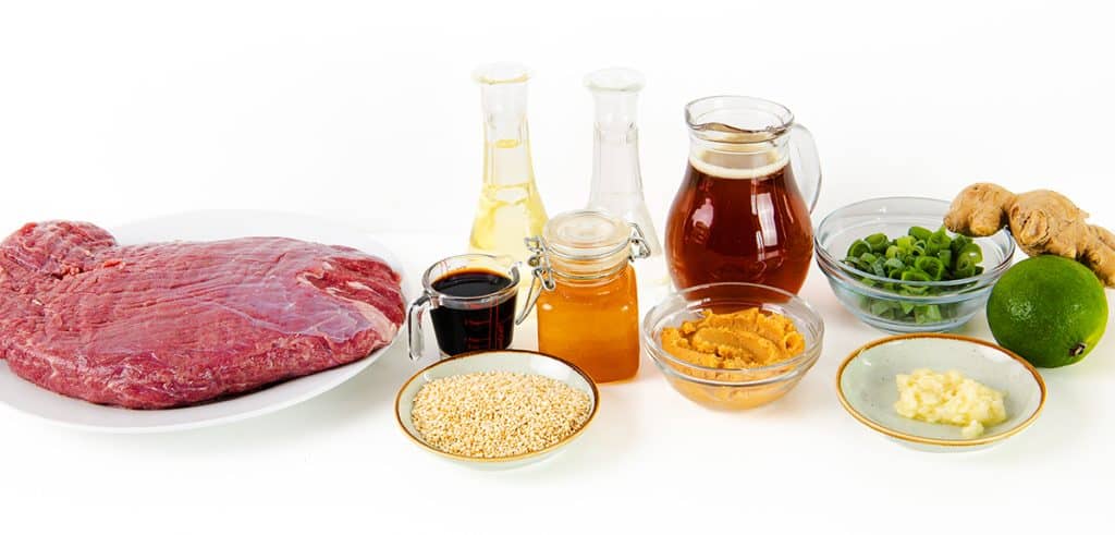 Raw beef on a plate, surrounded by sesame seeds, soy sauce, honey, peanut butter, miso paste, grated ginger, garlic cloves, lime, chopped green onions, and jars of oil and broth—all set against a white background. Perfect for crafting a flavorful glazed steak.