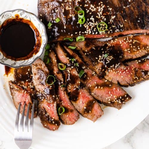 Sliced glazed steak topped with sesame seeds and green onions, served with a small bowl of dark miso sauce on a white plate. Hints of garlic elevate the flavors, while a fork rests next to the steak slices.