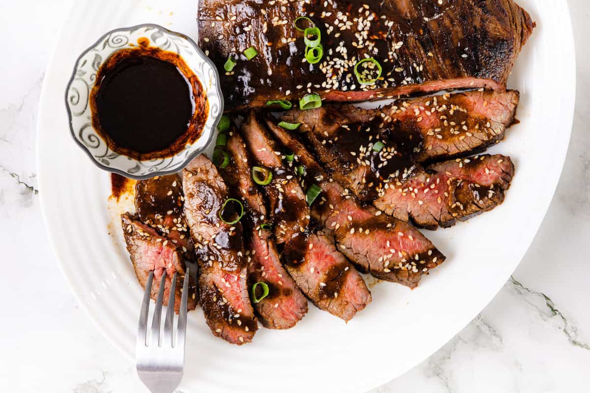 Sliced glazed steak topped with sesame seeds and green onions, served with a small bowl of dark miso sauce on a white plate. Hints of garlic elevate the flavors, while a fork rests next to the steak slices.
