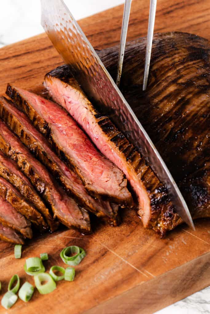 A knife slices through a juicy, grilled steak on a wooden cutting board, revealing a pink center. Sliced green onions and hints of savory garlic lie nearby.