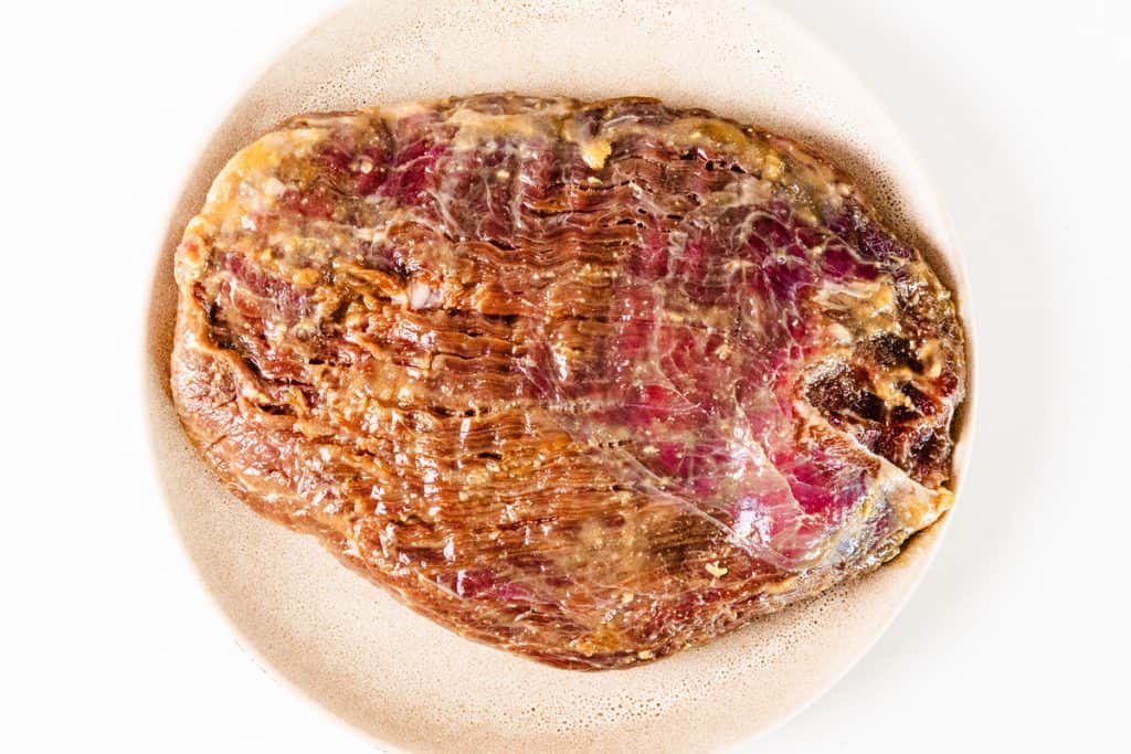 A raw corned beef brisket sits on a beige plate, showcasing its marbled meat and visible spices on the surface, with subtle hints of garlic enhancing its savory aroma.