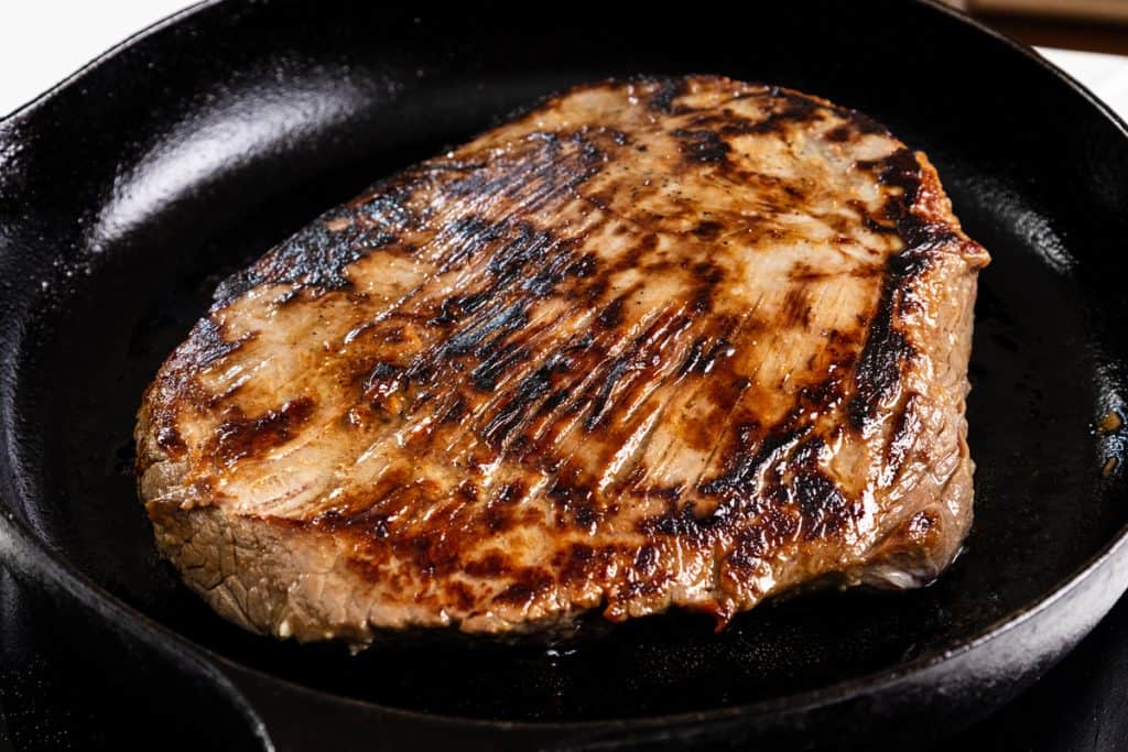 A thick, seared Garlic-Glazed Steak is cooking in a black cast iron skillet, showing char marks and a browned crust on the surface.