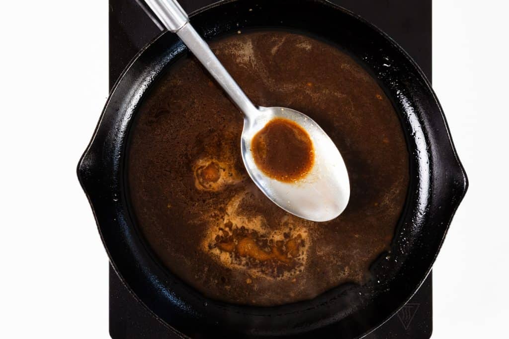 A metal spoon rests in a black pan containing a rich brown miso sauce or gravy on a stovetop. The spoon sits in the thick liquid, creating a light circle in the center of the pan—a perfect base for preparing glazed steak or infusing savory garlic flavor.