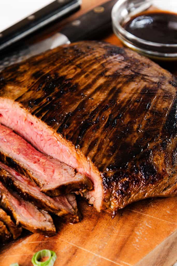 Sliced glazed steak with a juicy pink center on a wooden cutting board, accompanied by a bowl of rich garlic sauce and a knife in the background.
