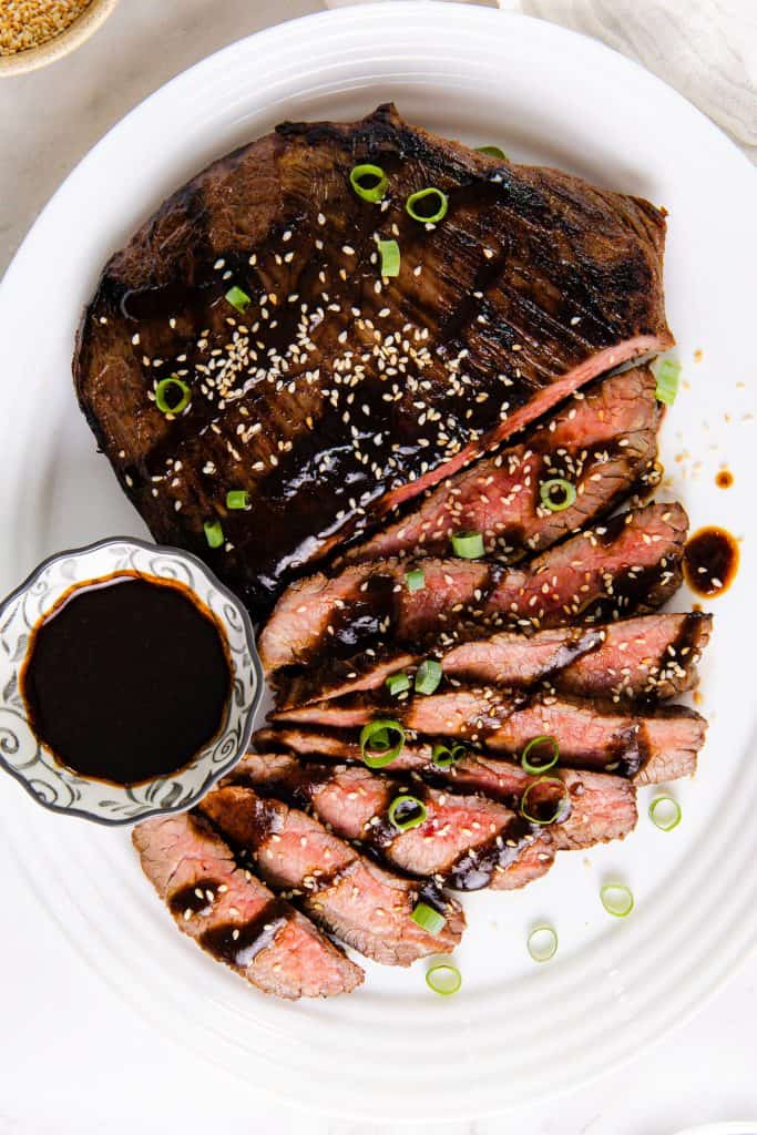 Sliced miso-glazed steak topped with sesame seeds and chopped green onions, served with a small bowl of dark garlic dipping sauce on a white plate.