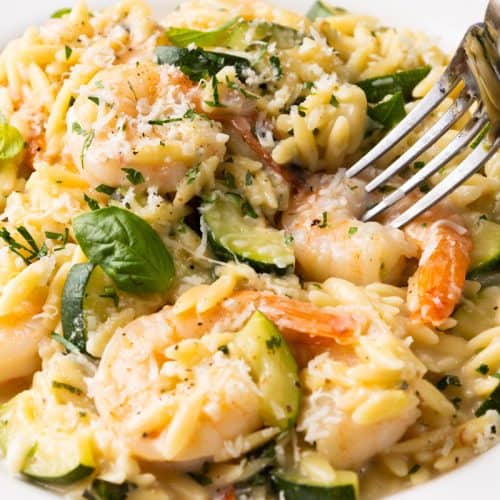 A close-up of creamy one-pan orzo pasta with succulent shrimp, zucchini, grated cheese, and herbs, garnished with fresh basil. A fork is poised to pick up some of the flavorful orzo and tender shrimp.