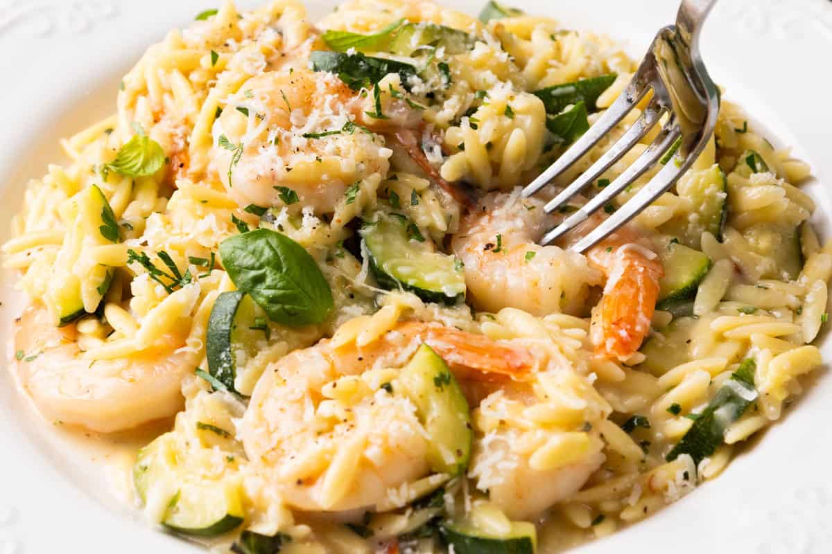 A close-up of creamy one-pan orzo pasta with succulent shrimp, zucchini, grated cheese, and herbs, garnished with fresh basil. A fork is poised to pick up some of the flavorful orzo and tender shrimp.