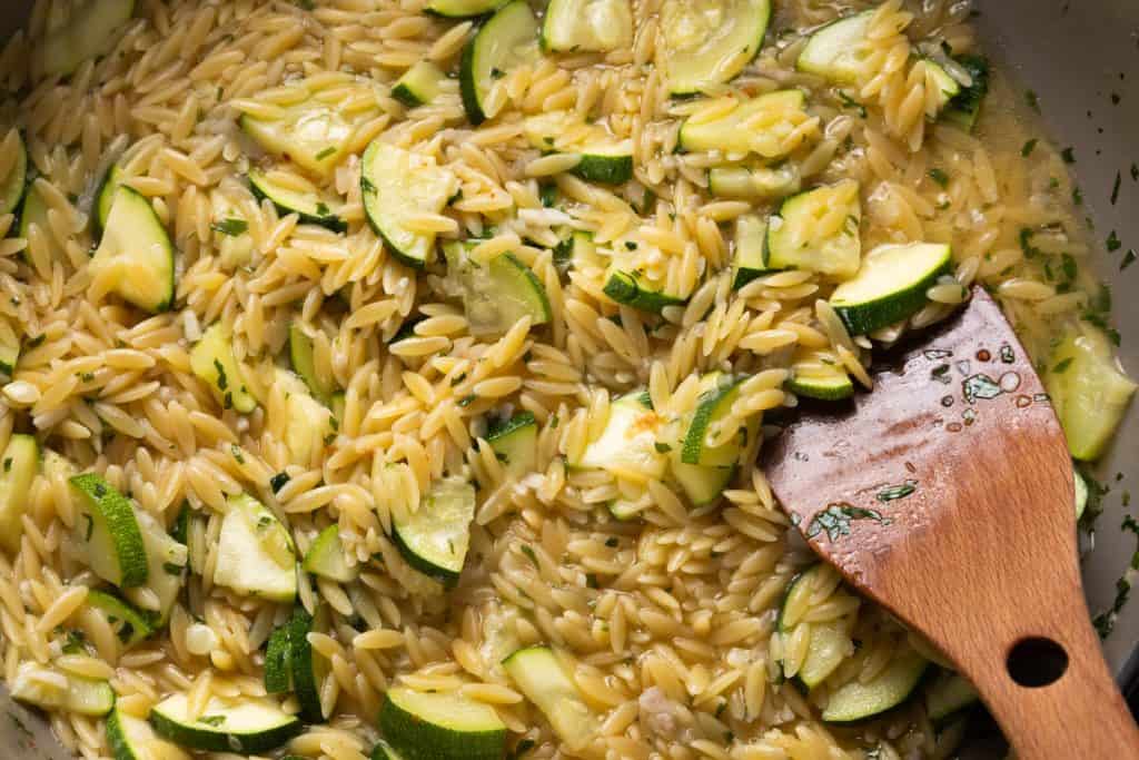 One-pan orzo pasta mixed with sliced zucchini in a pan, being stirred with a wooden spoon. The dish appears to be cooked in a light, broth-based sauce.