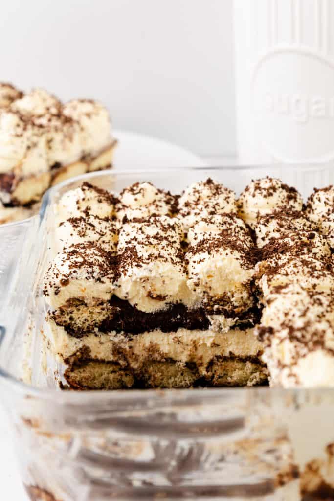 A glass dish of Hot Chocolate Tiramisu with a portion removed, revealing its creamy layers and chocolate topping. A slice is on a plate in the background, alongside a white sugar canister.