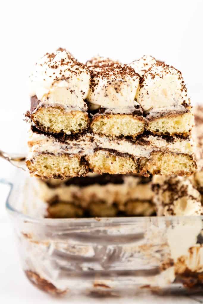 A close-up of a slice of Hot Chocolate Tiramisu with layers of coffee-soaked ladyfingers, creamy mascarpone, and cocoa powder, being lifted from a glass dish with a serving spatula.