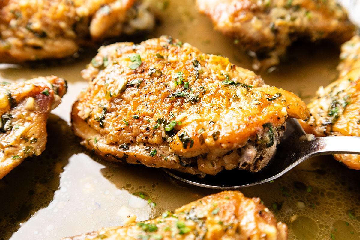 A close-up of a spoon lifting a piece of golden-brown, herb-seasoned chicken from a pan with flavorful juices and more chicken pieces.
