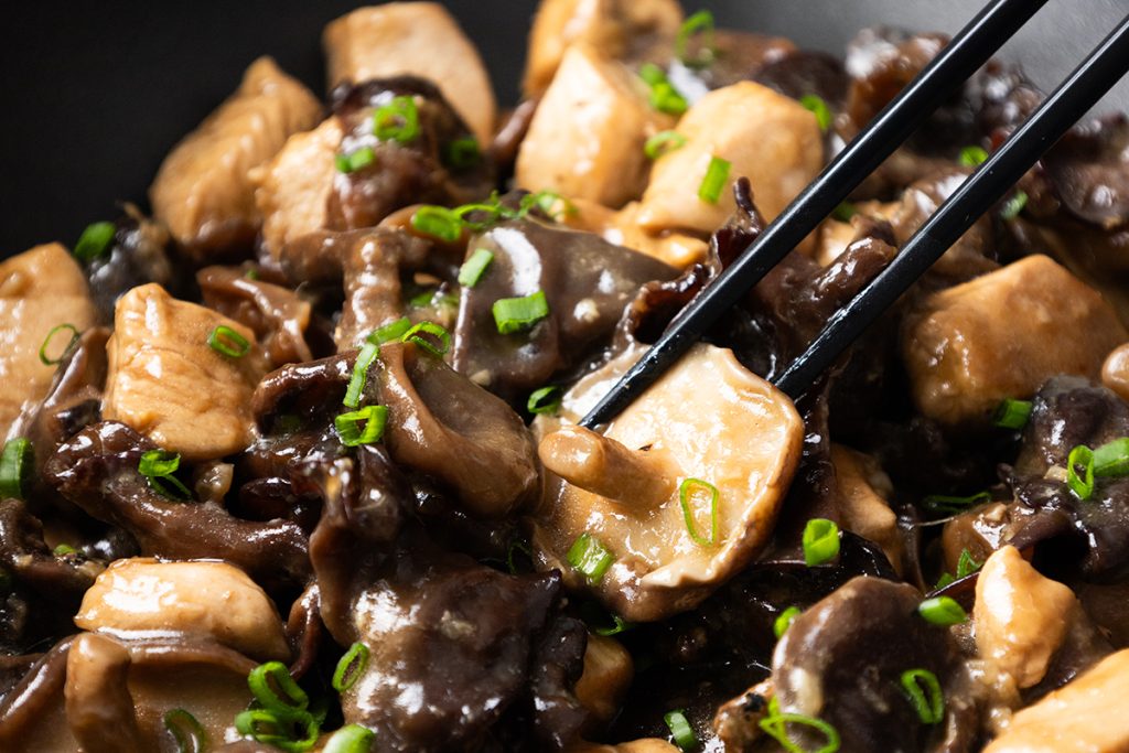 Close-up of chopsticks holding a piece of chicken with mushrooms in a glossy brown sauce, garnished with chopped green onions.
