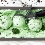 Rectangular container of Mint Chocolate Chip ice cream with three scooped balls, a metal ice cream scooper, mint leaves, and chocolate chunks on top and around the ice cream.