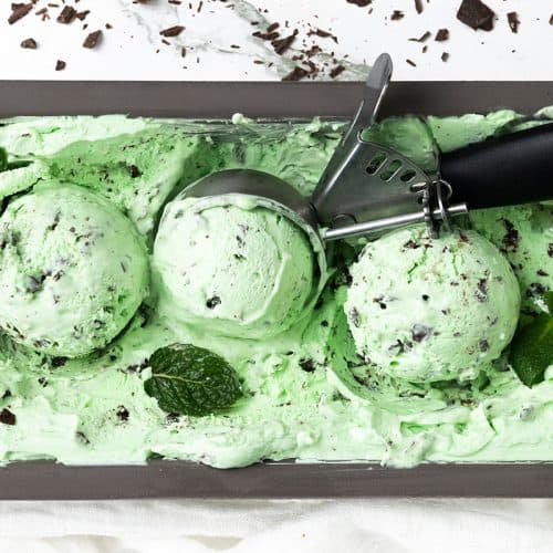 Rectangular container of Mint Chocolate Chip ice cream with three scooped balls, a metal ice cream scooper, mint leaves, and chocolate chunks on top and around the ice cream.