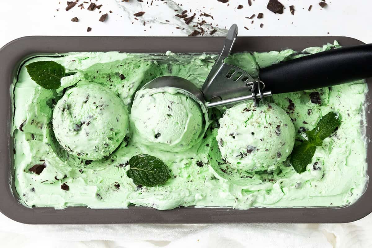 Rectangular container of Mint Chocolate Chip ice cream with three scooped balls, a metal ice cream scooper, mint leaves, and chocolate chunks on top and around the ice cream.