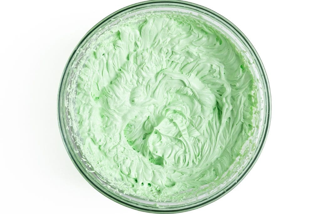 A glass bowl filled with fluffy, light green whipped frosting or cream, reminiscent of mint chocolate chip ice cream, viewed from above on a white background.