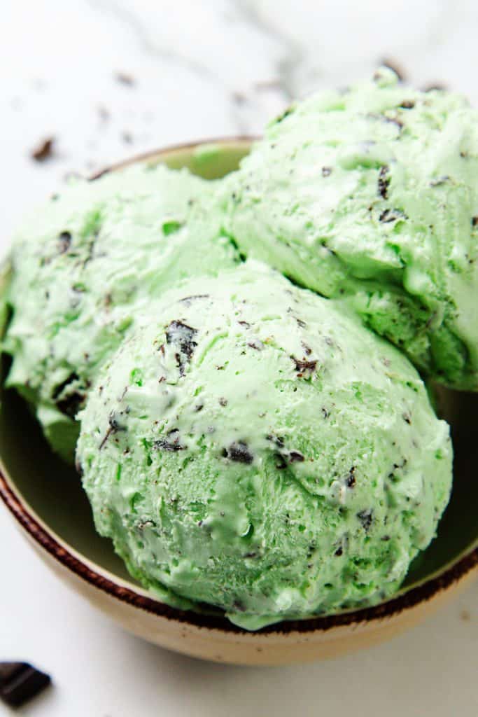 Three scoops of fresh mint ice cream with chocolate chips sit in a brown bowl, their creamy green color accentuated by the visible flecks of chocolate, all placed on a pristine white surface.