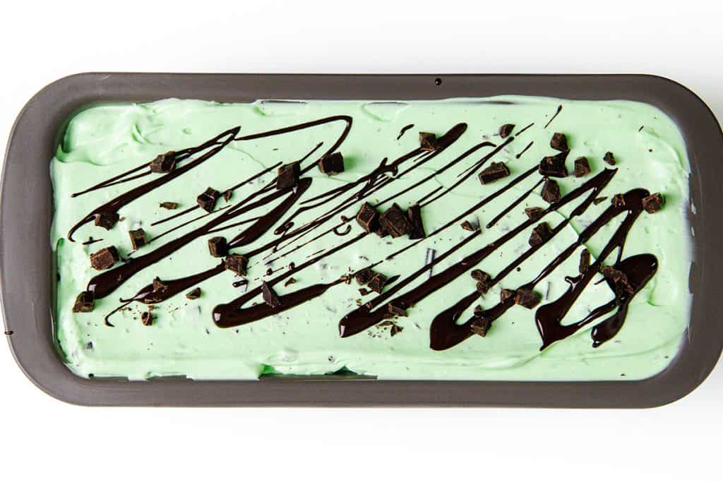 Rectangular pan filled with creamy mint chocolate chip ice cream, topped with a drizzle of chocolate syrup and generous chocolate chunks, on a white background.
