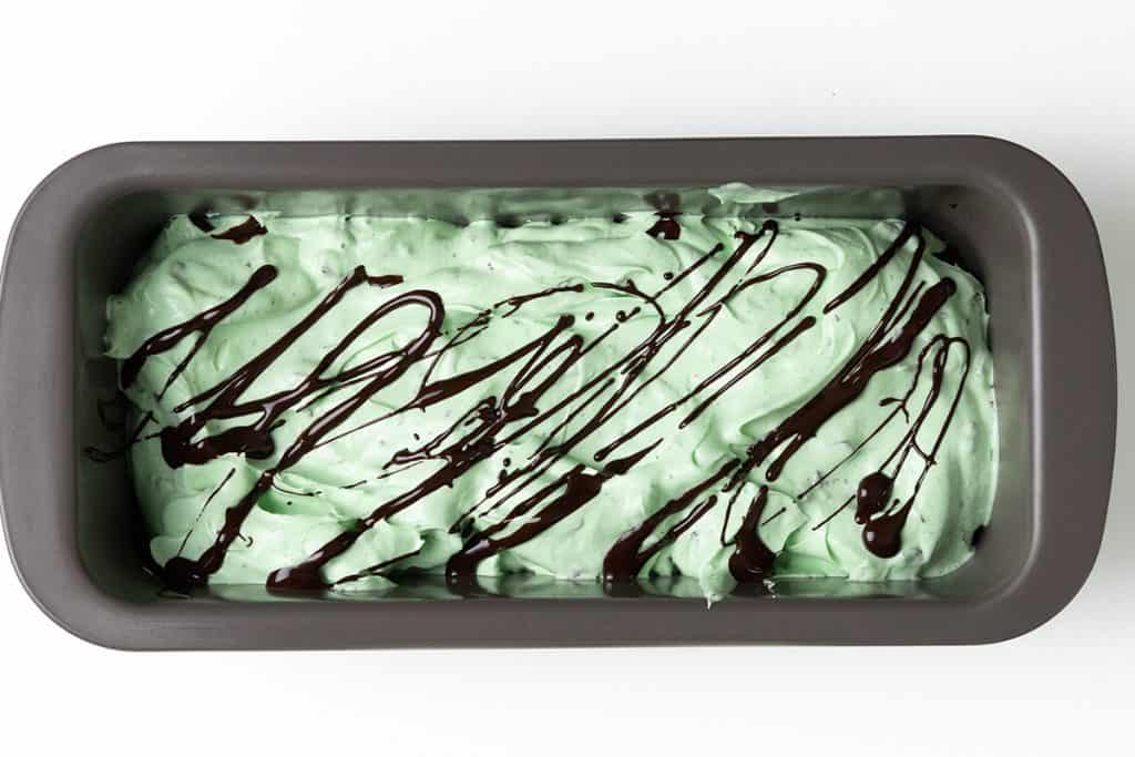 Rectangular pan filled with light green mint chocolate chip ice cream, topped with drizzled chocolate syrup, viewed from above on a white background.