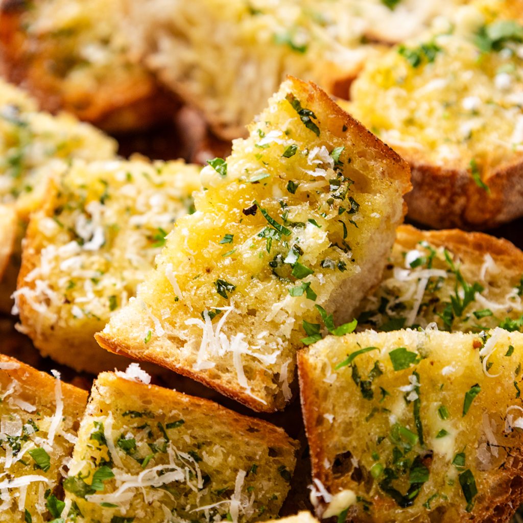 Slices of garlic bread topped with melted butter, chopped parsley, and grated cheese, stacked closely together.