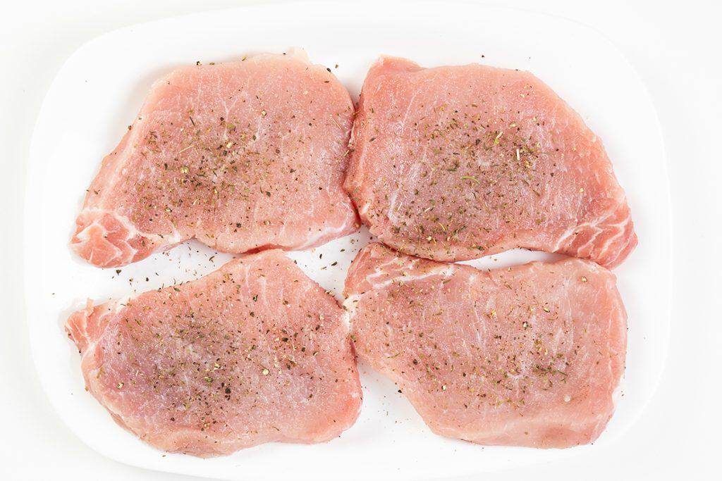 Four raw Mediterranean-style pork chops seasoned with ground pepper and herbs, arranged on a white rectangular plate against a white background.