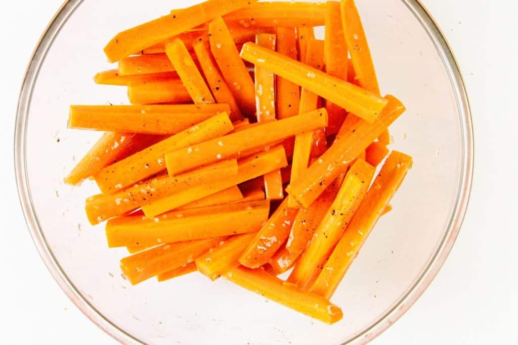 Carrot sticks in a clear glass bowl, sprinkled with coarse salt and black pepper, evoke a fresh Mediterranean flair on a white background.