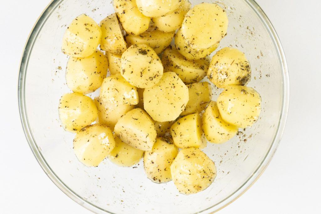 A glass bowl filled with peeled, sliced potatoes seasoned with Mediterranean herbs and spices, viewed from above on a white background.