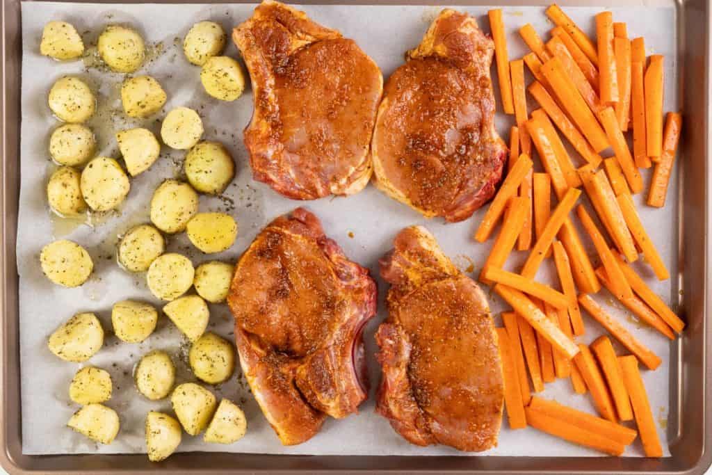 A Mediterranean sheet pan showcases four seasoned pork chops, sliced carrots, and halved baby potatoes, all arranged on parchment paper and ready to be cooked.
