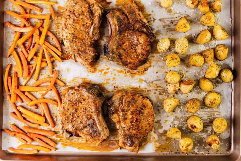 Sheet pan with Mediterranean-inspired roasted pork chops in the center, sliced carrots on the left, and halved baby potatoes on the right, all perfectly seasoned and baked for a flavorful meal.