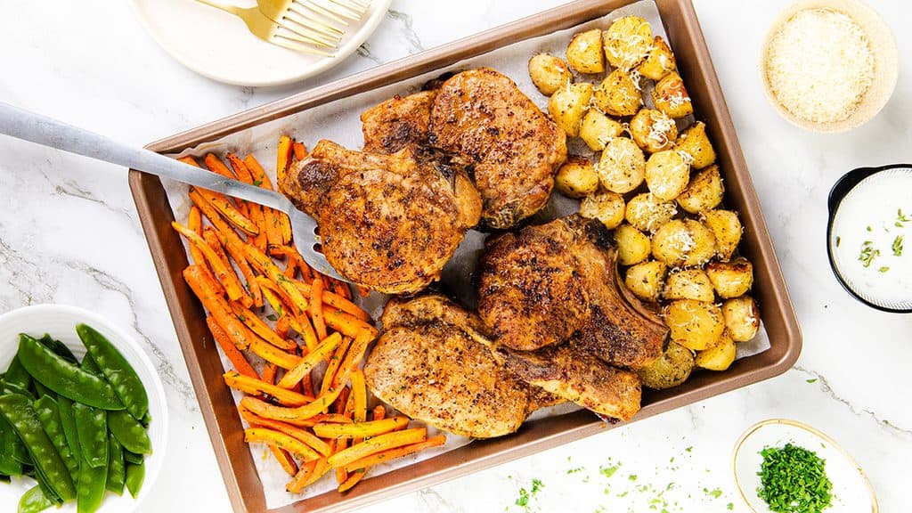 A Mediterranean-inspired sheet pan meal featuring four seasoned pork chops, roasted baby potatoes topped with grated cheese, and roasted carrot sticks. Nearby bowls hold snap peas, sauce, shredded cheese, and chopped herbs on a white surface.