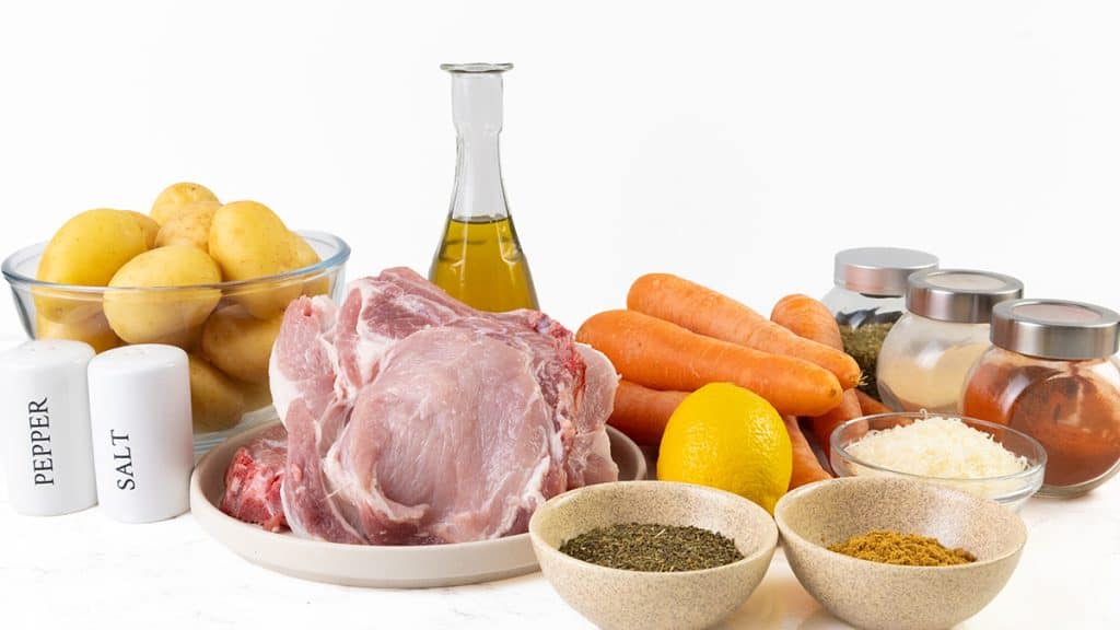 A collection of cooking ingredients perfect for a Mediterranean-inspired meal: raw pork chops, potatoes, carrots, a lemon, oil, salt, pepper, dried herbs, grated cheese, and spice jars—all thoughtfully arranged on a white surface and ready for your next flavorful sheet pan creation.