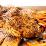A close-up of seasoned, grilled pork chops on a sheet pan, served with roasted carrot sticks and potatoes. A metal spatula is lifting one chop. The food is golden brown and looks freshly cooked, with Mediterranean-inspired flavors throughout.