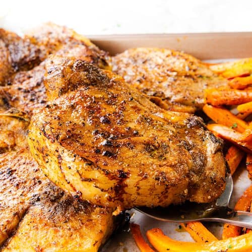 A close-up of seasoned, grilled pork chops on a sheet pan, served with roasted carrot sticks and potatoes. A metal spatula is lifting one chop. The food is golden brown and looks freshly cooked, with Mediterranean-inspired flavors throughout.
