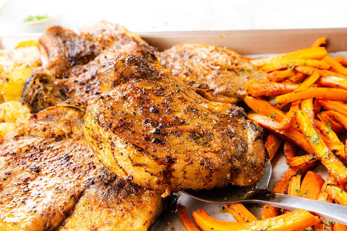A close-up of seasoned, grilled pork chops on a sheet pan, served with roasted carrot sticks and potatoes. A metal spatula is lifting one chop. The food is golden brown and looks freshly cooked, with Mediterranean-inspired flavors throughout.
