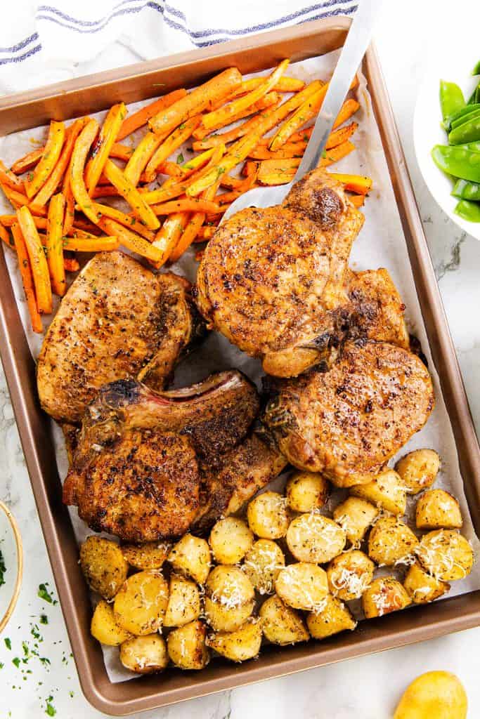 A Mediterranean-inspired baking sheet with juicy pork chops, seasoned potato halves topped with herbs and cheese, and roasted carrot sticks. A spatula rests on the tray, while vibrant vegetables are visible on the side—perfect for an easy sheet pan pork chops dinner.